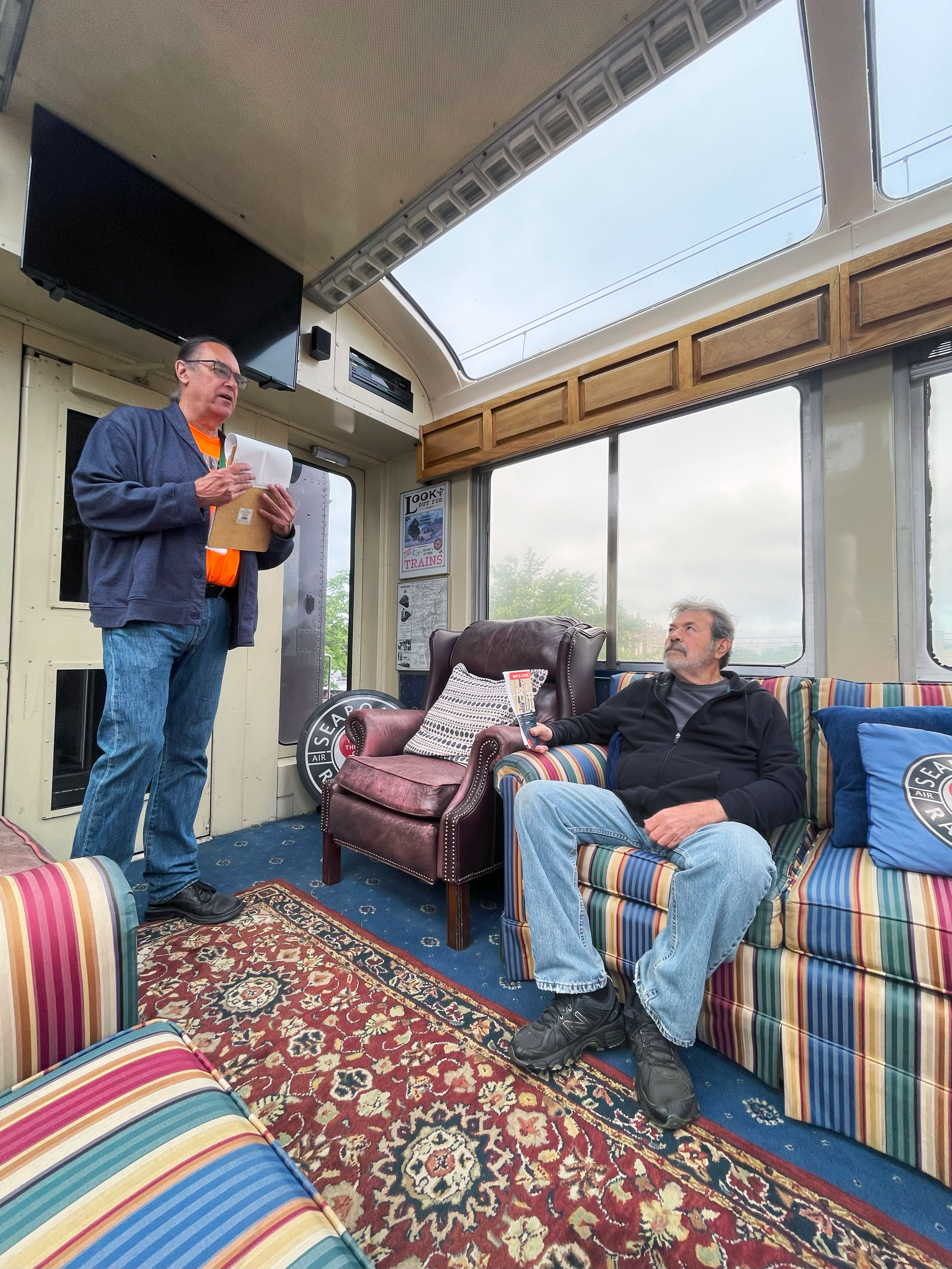a man sitting on a couch on a pullman car with another man speaking to him